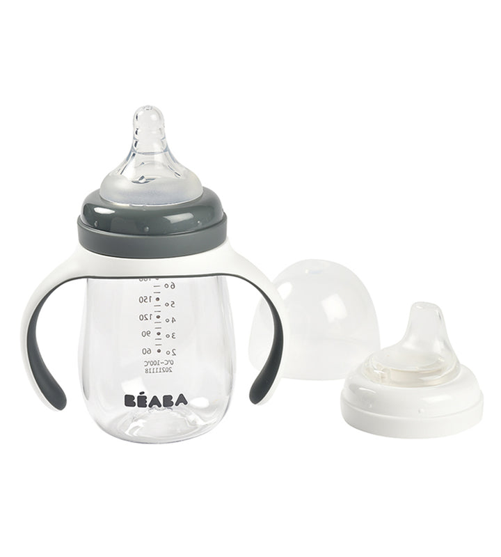 Beaba 2 In 1 Learning Bottle set including a transparent bottle with blue handles and measurement markings, a bottle teat, and a spout with two rings.