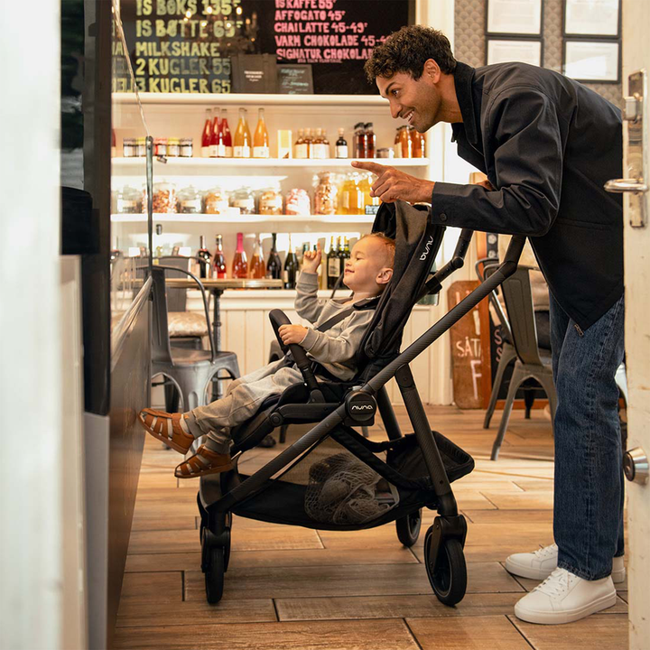 Man pushing a stroller with a child inside, both looking at a menu board in a cafe.